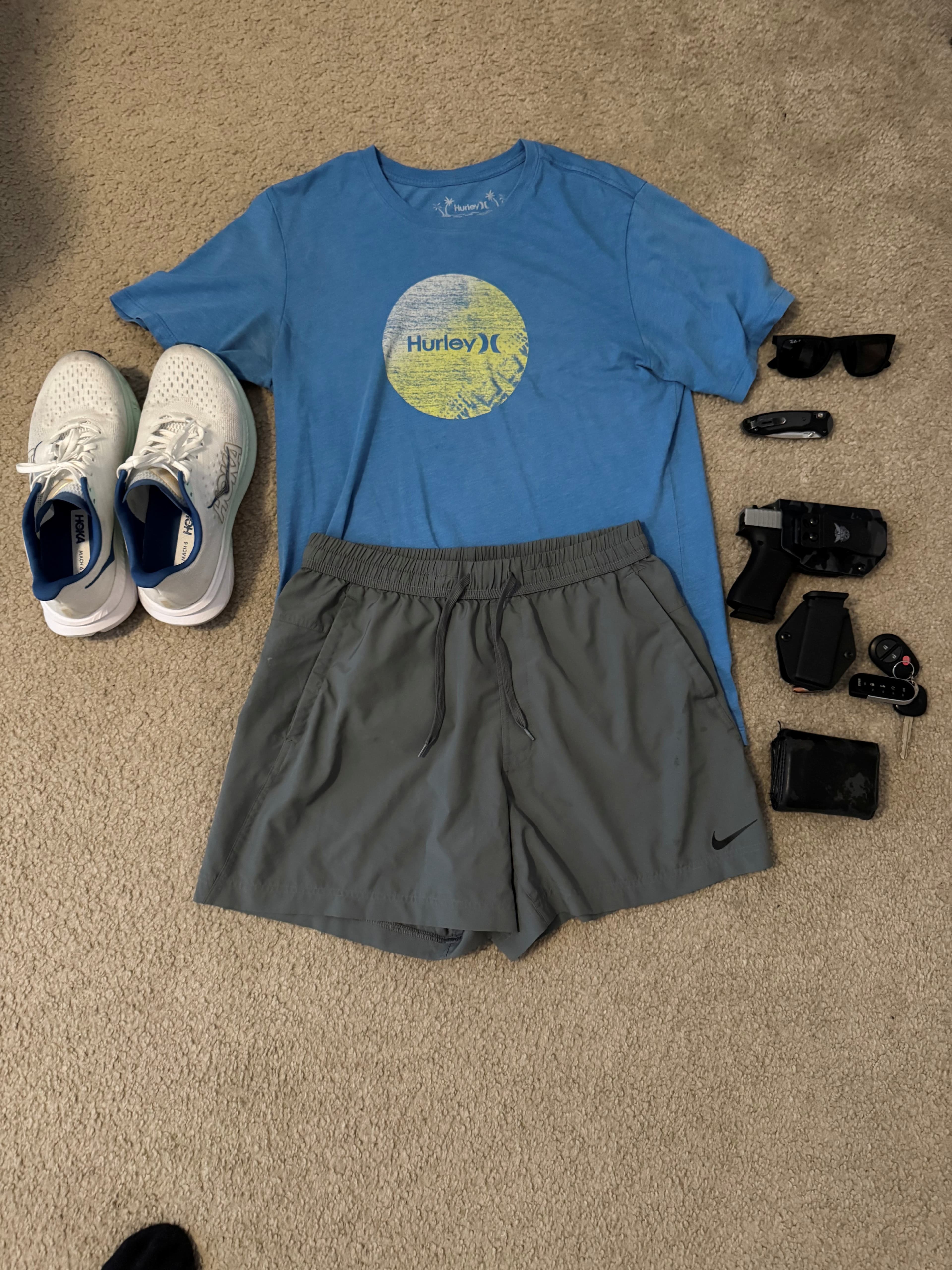 Everyday carry items including wallet, Glock 43X, pocket knife, keys, sunglasses, hat, and phone laid out on clothing